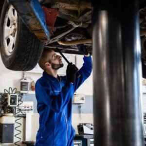 Reparación Coches Málaga Arenas. Mechanic in blue coveralls inspecting car undercarriage on hydraulic lift.